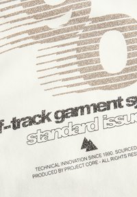 De tekst op de witte stof zegt "track garment standard issue" met verspreide tan kleurige horizontale lijnen en een klein berglogo eronder.