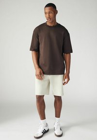 T-shirt oversize marron en coton, associé à un short crème clair. Des baskets blanches avec des rayures noires et des chaussettes blanches complètent le look.