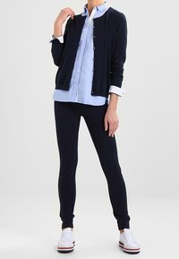 Cardigan blu navy sopra una camicia azzurra a bottoni, abbinato a leggings blu navy e sneaker bianche. Tessuto liscio, design aderente e dettagli sui bottoni discreti.