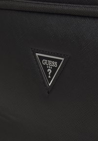 Punto in materiale nero testurizzato con un logo triangolare argento con la scritta "GUESS USA" e un punto interrogativo, su una superficie nera liscia.