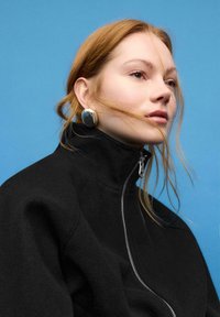 Sort fleecejakke med høj krave og sølvfarvede lynlåse. Modellen har store sølvfarvede øreringe og står foran en ensartet blå baggrund.