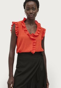 Rote Rüsche-Bluse mit tiefem V-Ausschnitt und kurzen Ärmeln, kombiniert mit einem schwarzen Wickelrock. Der Stoff hat eine strukturierte Oberfläche.