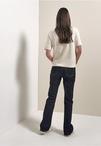 Beige short-sleeve t-shirt, dark blue flared jeans, and white shoes, viewed from the back against a neutral backdrop.