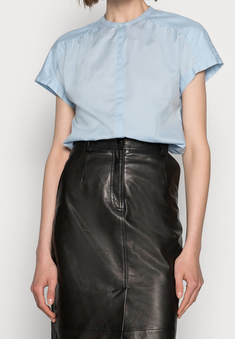 Personne portant un chemisier bleu clair à manches courtes rentré dans une jupe en cuir noire taille haute sur un fond uni.