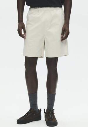 Mand iført beige shorts med elastisk talje, mørke sneakers med grå mellemlange sokker, stående mod en ensfarvet baggrund.