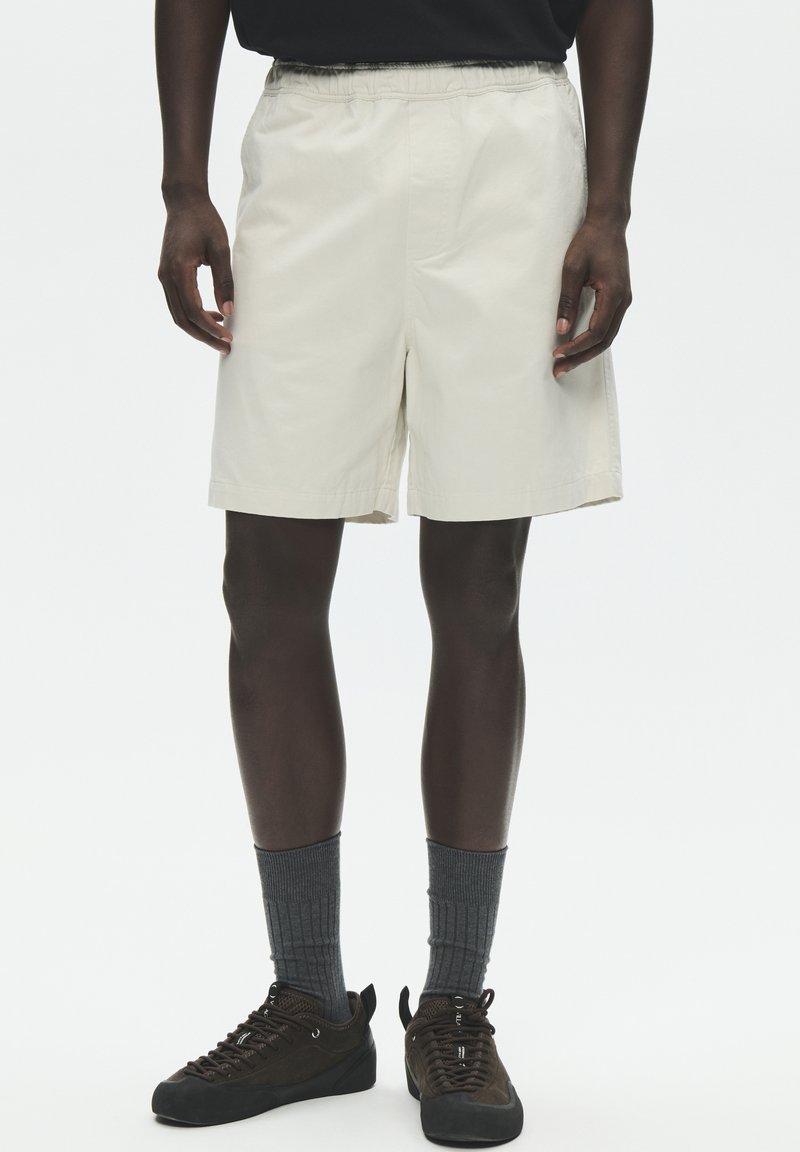 Homme portant un short beige à taille élastique, des baskets sombres avec des chaussettes grises montant jusqu'au mollet, debout devant un fond uni.