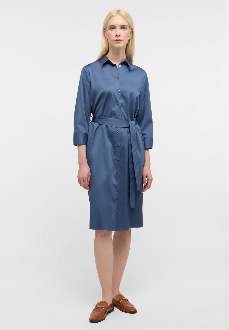 Blaues Hemdkleid mit langen Ärmeln aus einem glatten Stoff, mit Kragen, vorderem Knopfverschluss und einem gebundenen Taillengürtel. Braune Loafers runden den Look ab.