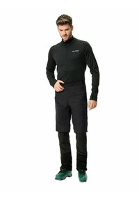 Maglia nera da uomo con zip a tre quarti a maniche lunghe abbinata a pantaloni corti neri isolati fino al ginocchio. Presenta stivali da sci verde scuro e una texture liscia.