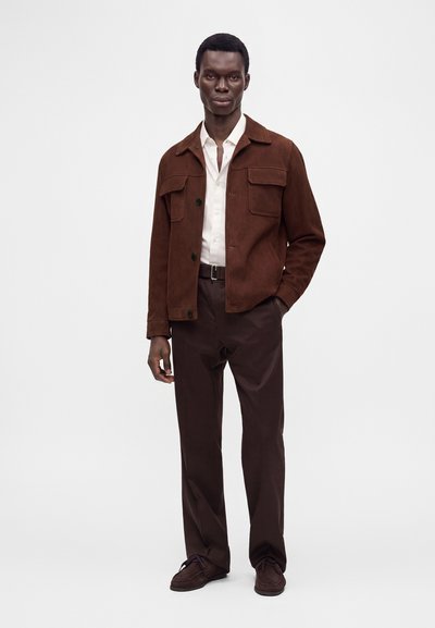 Homme portant une veste marron par-dessus une chemise blanche, un pantalon brun foncé et des chaussures marron, debout devant un fond clair uni.