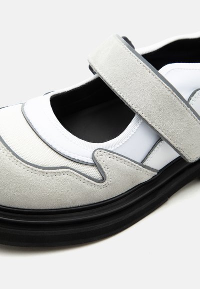 Chaussure Mary Jane blanche et grise avec bride en daim, semelle noire rembourrée et tige en tissu blanc élastique.