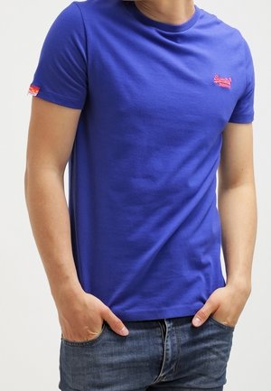 Lilla bomuld t-shirt med korte ærmer, rund hals og et lille rødt logo på øverste venstre side. Bruges med denim jeans.