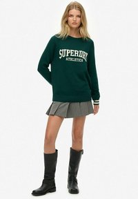 Groen gebreide trui met de tekst "SUPERDRY ATHLETICS", gecombineerd met een grijze geplooide rok met fijne strepen en zwarte kniehoge laarzen.