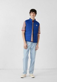 Joven de pie con chaleco acolchado azul, camiseta de manga corta a rayas, jeans azul claro y zapatillas blancas, frente a una pared blanca.