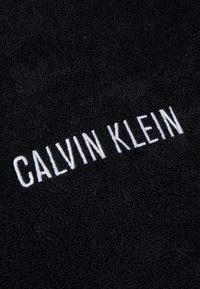 Črn brisača z teksturirano tkanino in obrobljeno površino. "CALVIN KLEIN" izvezen z belo nitjo, centrirano in poudarjeno.
