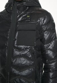 Veste en duvet noire avec une texture matelassée brillante, capuche, fermeture éclair à l'avant et plusieurs poches zippées et à rabat. Présente des accents de logo.