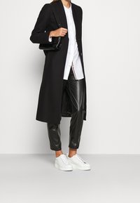 Abrigo largo negro sobre una camisa blanca de botones, combinado con pantalones cortos negros brillantes y zapatillas de plataforma blancas. Pequeño bolso de mano negro.
