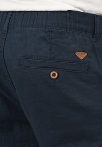 Mörkblå linneblandade shorts med en bakficka med knappstängning och en läderliknande märkesetikett formad som en triangel.