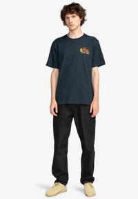 Navy blue cotton t-shirt with short sleeves, featuring a yellow graphic print on the left chest. Paired with dark denim jeans and beige shoes.