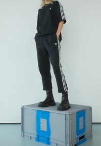 Černé oversized tričko a sportovní kalhoty s bílými bočními pruhy, zkombinované s robustními černými botami, vystavené na šedé krabici.