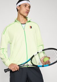 Φούξια φούτερ με φερμουάρ σε νέον πράσινο χρώμα, με αντίθετες ραφές και λογότυπο. Μαύρη ρακέτα τένις κρατιέται με μια μπάλα τένις στο χέρι.