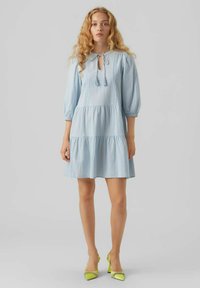 Robe en coton bleu clair avec des manches bouffantes trois-quarts, un col noué et une jupe à volants. Assortie à des escarpins verts fluo.