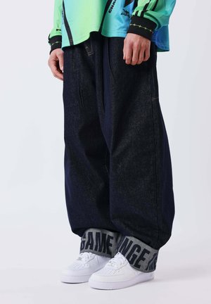 Persona que lleva vaqueros oscuros de pierna ancha con grandes puños grises con la etiqueta "GAME" y zapatillas blancas, combinados con una camiseta de manga larga verde y azul.