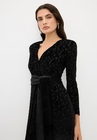 Schwarzes Samtkleid mit langen Ärmeln, tiefem Ausschnitt, Blumenmuster und einem Stoffgürtel, der an der Taille gebunden ist. Sanfte Textur, elegantes Design.