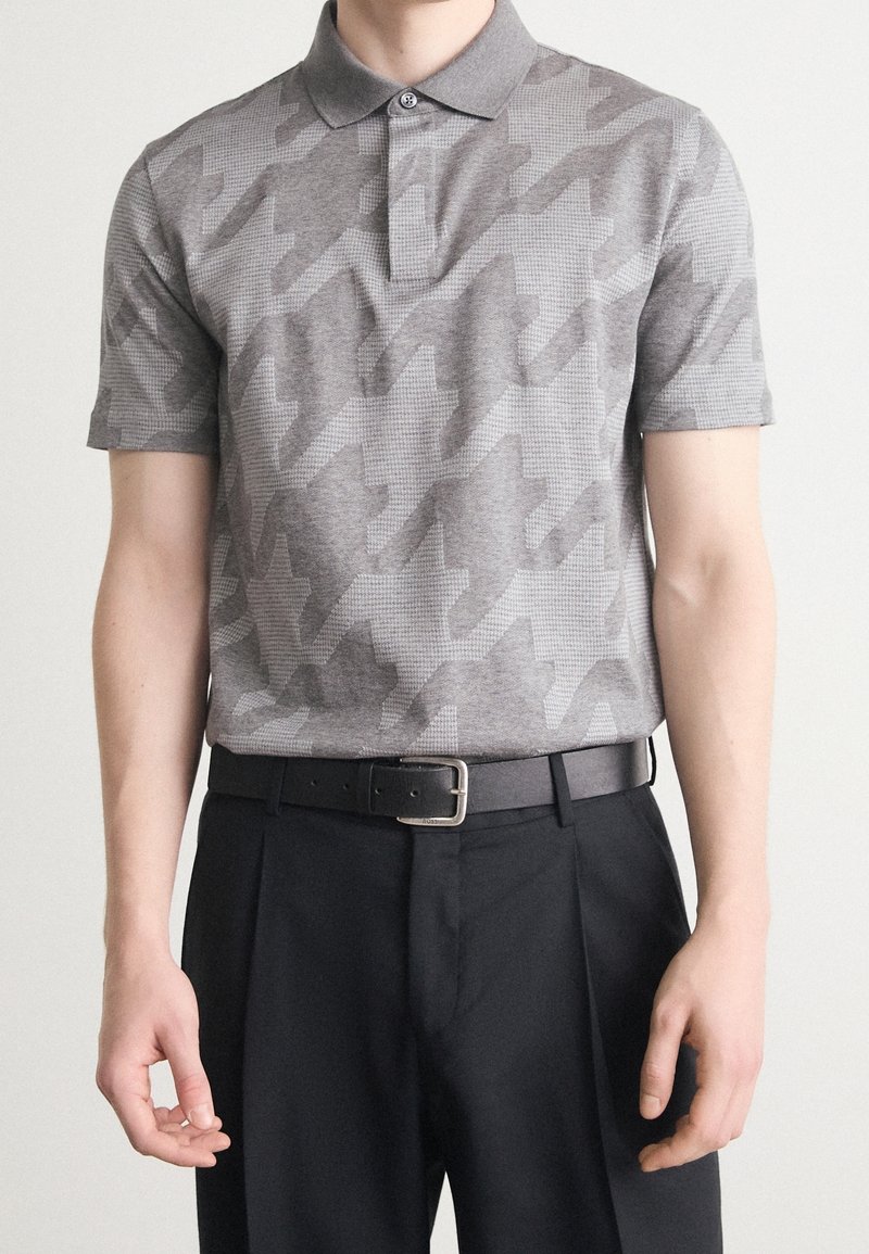 Homme portant un polo gris à motifs, rentré dans un pantalon noir avec une ceinture en cuir noir et une boucle argentée, debout devant un fond clair.