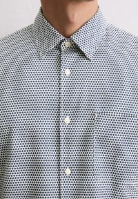 Overhemd met knopen in een blauw-wit geometrisch patroon, met een puntige kraag en een borstzak, gemaakt van een gestructureerde stof.