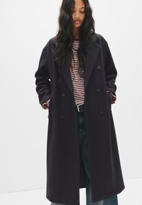 Donna con lunghi capelli scuri che indossa un cappotto doppiopetto scuro sopra una camicia a righe rossa e bianca e jeans blu, con le mani nelle tasche del cappotto.