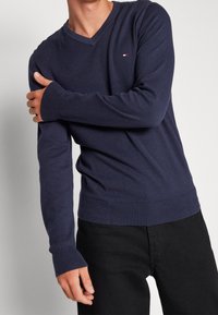 Pull col en V bleu marine en tissu tricoté doux. Comprend des poignets et un ourlet côtelés. Petit logo discret sur la poitrine.