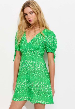 Młoda kobieta z długimi blond włosami, ubrana w zieloną krótką sukienkę w kwiaty z bufkami na rękawach i dekoltem w serek, stojąca na białym tle.