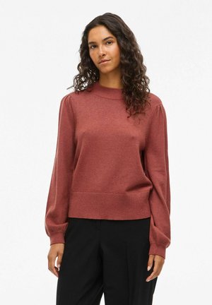 Maglione rosso con collo alto e maniche a palloncino. Realizzato in un materiale morbido, presenta un orlo a coste e una vestibilità rilassata. Indossato con pantaloni neri.