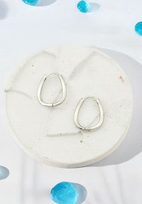 Silver hoops med tårform, visade på en strukturerad betongskål, omgiven av blå dekorativa stenar.