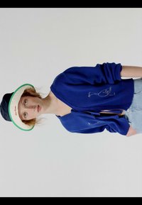 Joven mujer con un cárdigan azul con un bordado de pájaro, shorts de mezclilla claros y un sombrero estilo bucket blanco y negro con borde verde.