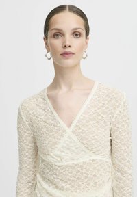 Top in pizzo bianco con design avvolgente, dotato di scollatura a V, maniche lunghe e dettagli di bordatura delicata. Il pizzo presenta un motivo floreale.
