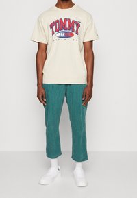 Béžové bavlněné tričko s červeným a modrým logem "TOMMY JEANS ATHLETICS", spárované s tyrkysovými kordovými kalhotami a bílými teniskami.