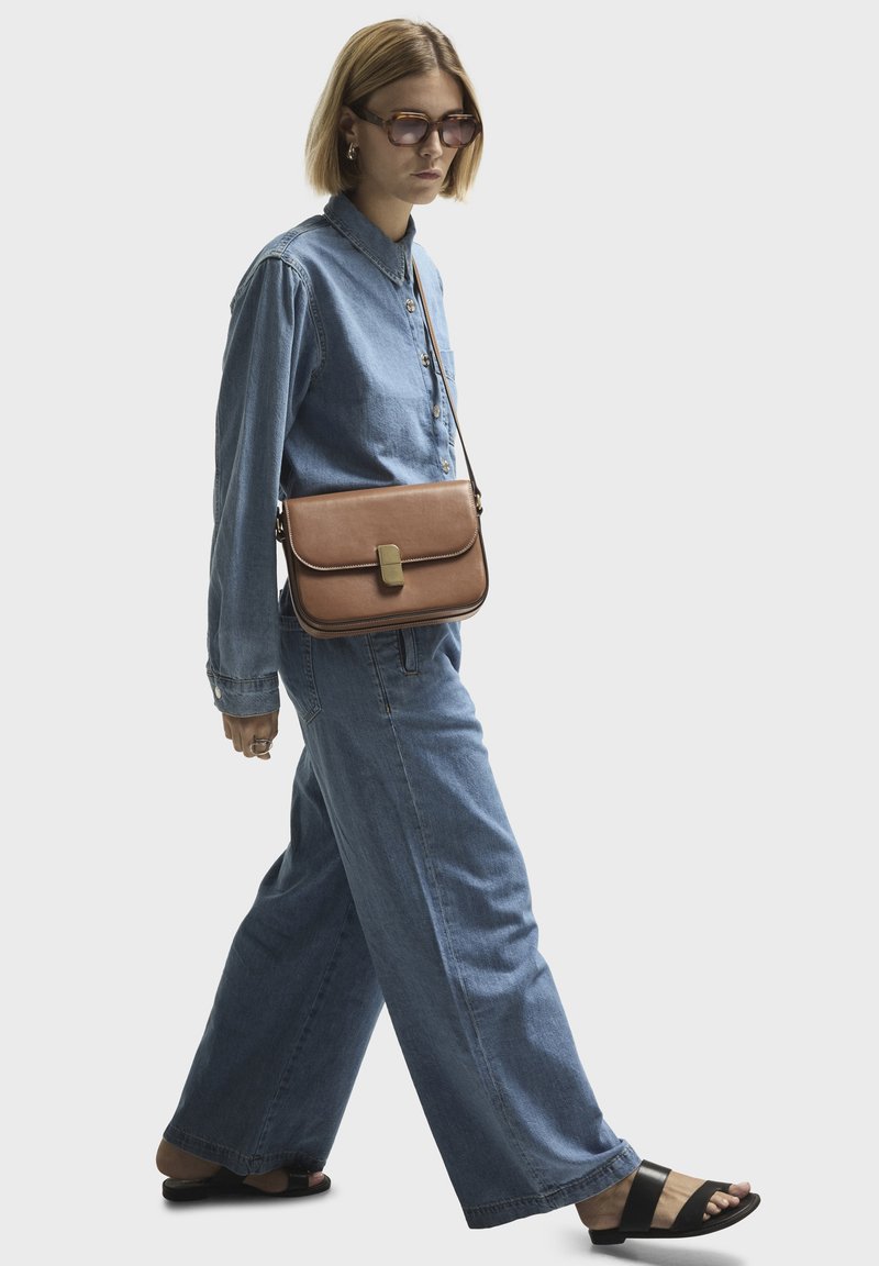Brown leather crossbody bag with a flap and gold-tone clasp, paired with a denim shirt and wide-leg jeans, complemented by black sandals.