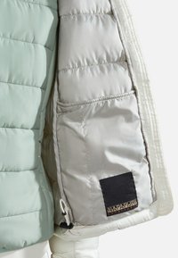 Oc cieplana kurtka z jasnooliwkowym zewnętrzem, srebrnym wnętrzem i czarną łatą z logo marki. Posiada pikowaną teksturę oraz kieszenie zapinane na zamek.