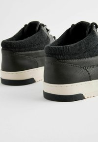 Zapatillas negras de cuero con un cuello de tejido texturizado, media suela blanca y suela negra. Presenta herrajes metálicos y detalles de costura mínima.