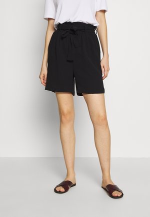 Kvinde iført sorte shorts med høj talje og bindebånd foran, en hvid t-shirt og mørkebrune åbne slide-sandaler, stående på en ensfarvet baggrund.