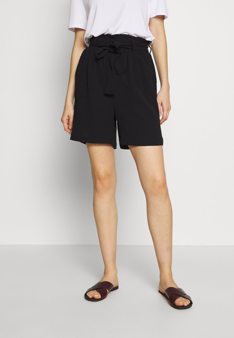 Femme portant un short noir taille haute avec un nœud à l’avant, un t-shirt blanc et des sandales ouvertes marron foncé, debout sur un fond uni.