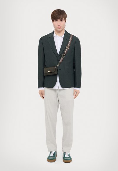 Sac à bandoulière noir avec une fermeture dorée, porté sur un blazer vert foncé, une chemise blanche, un pantalon beige et des baskets vertes avec des lacets blancs.