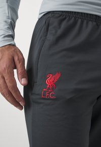 Pantalones atléticos de color gris oscuro con bolsillos laterales y un logo bordado en rojo del Liverpool FC en el muslo izquierdo. Textura de tela suave.