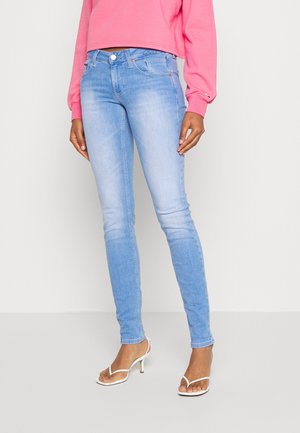 Mujer vestida con jeans ajustados de color azul claro, acompañados de sandalias de tacón blancas y una blusa corta de manga larga rosa, todo sobre un fondo neutro.