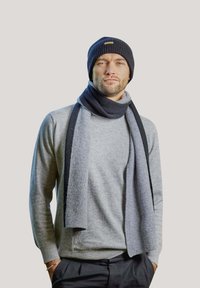 Set di berretto lavorato a maglia e sciarpa in grigio scuro e nero, indossato con un dolcevita grigio chiaro. Tessuto texturizzato con motivi a costine e un logo decorativo.