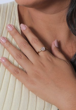 Gold ring with a round, light pink stone, worn on a hand resting against a textured beige fabric. Smooth finish and slender band.