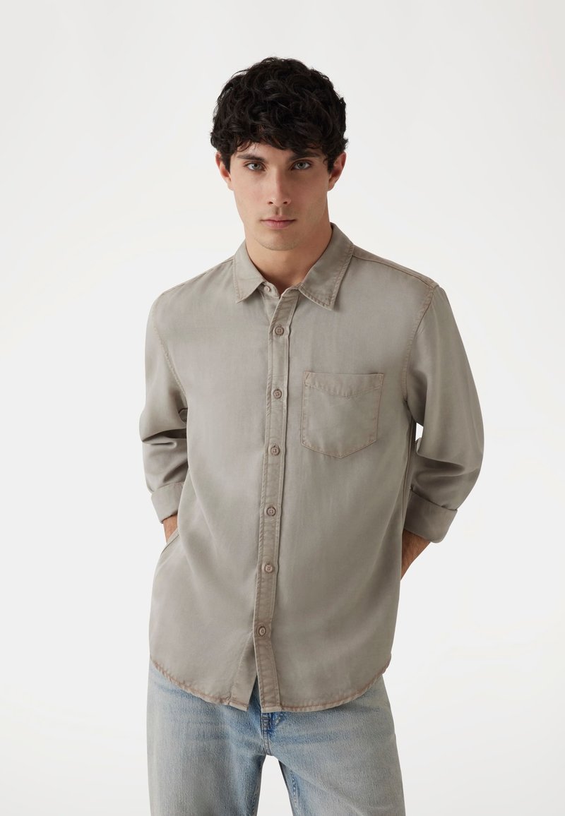 Camisa de botón gris claro con un bolsillo en el pecho, mangas largas arremangadas hasta los codos, y una textura de tela suave. Diseño casual con un dobladillo redondeado.