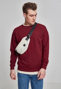 Rød hettegenser med lange ermer, brukt over en kremfarget topp, paret med blå jeans. En kremfarget crossover-veske med en svart stropp hviler på overkroppen.