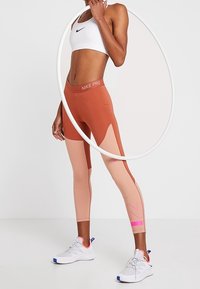 Legíny Nike Pro v terakotové a růžové barvě, se širokým pasem, kontrastními panely a logem písmene "N". Kombinováno s bílým sportovním podprsenkou.